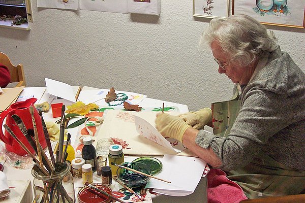 Bewohnerin beim Malen an einem Tisch voller Pinsel, Farbtöpfe und Papier
