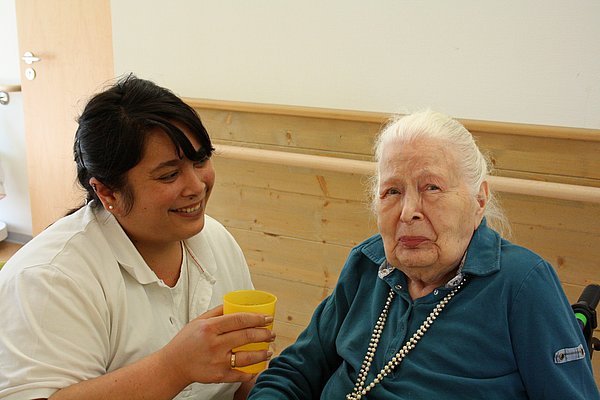 Bewohnerin neben einer Mitarbeiterin, die ihr zu Trinken gibt