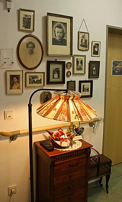 Wand im Flur des Haus am Urselbach mit persönlichen Bildern der Bewohner, davor eine Kommode und eine Stehlampe, sowie ein Nähkästchen
